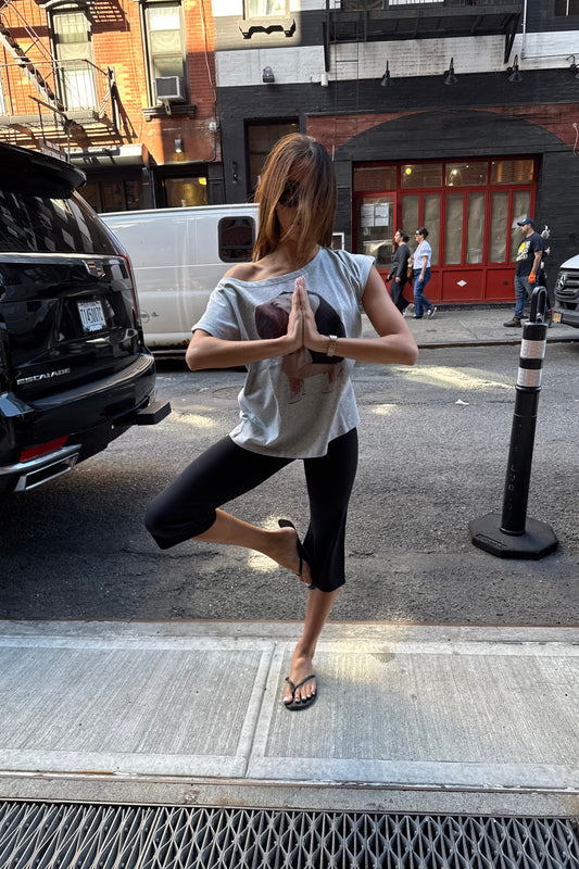 A woman in a gray graphic t-shirt and black capri leggings balances in a tree pose on a city street sidewalk, with parked cars and buildings in the background.