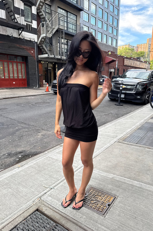 A woman wearing a black strapless dress stands on a city sidewalk next to a street with parked cars and buildings in the background.