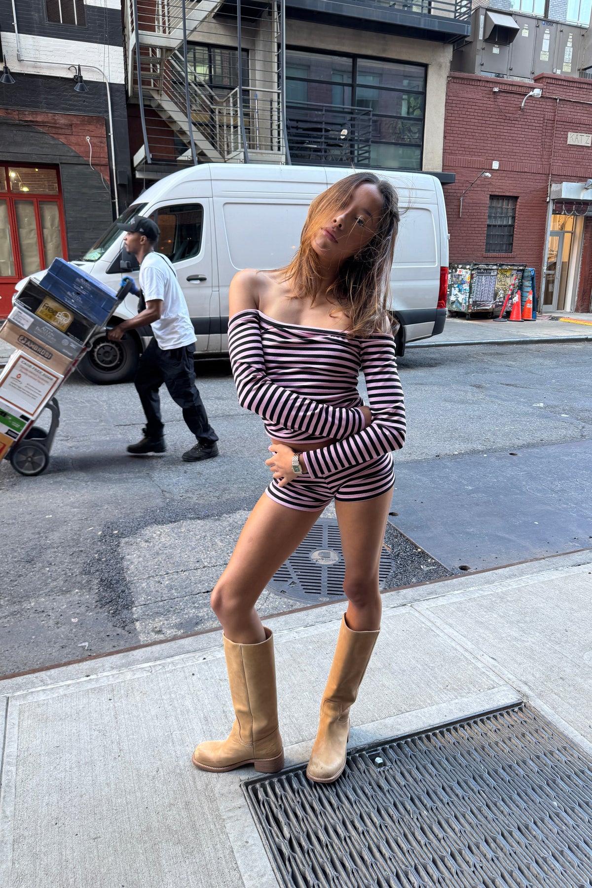 A woman posing on a city street wearing a matching off-the-shoulder long-sleeve top and shorts set in a pink and black striped pattern, paired with tan knee-high boots.
