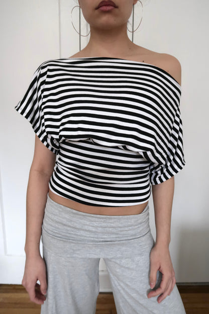 Person wearing a black and white striped off-shoulder top and gray pants against a plain background