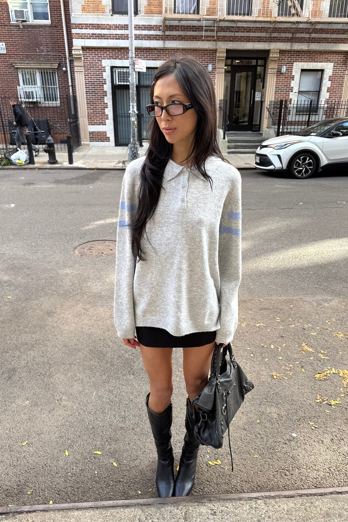 Woman wearing grey sweater with blue stripes standing on a street with a building and car in the background