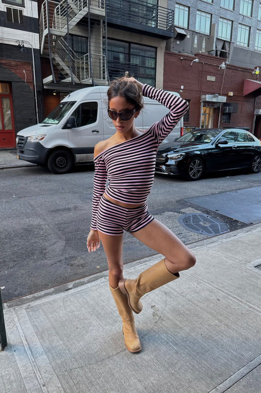 A woman posing on a city sidewalk wearing a matching two-piece set with a pink and black horizontal stripe pattern, consisting of an off-the-shoulder long-sleeve crop top and matching shorts. She is also wearing tan knee-high boots and sunglasses, with parked cars and urban buildings in the background.