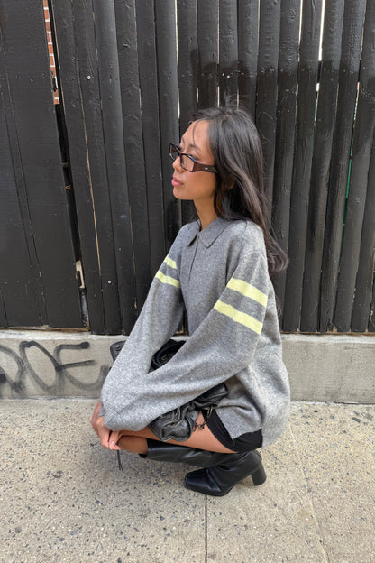 Person leaning against a wall wearing a grey sweater with a closed shutter on a sidewalk.