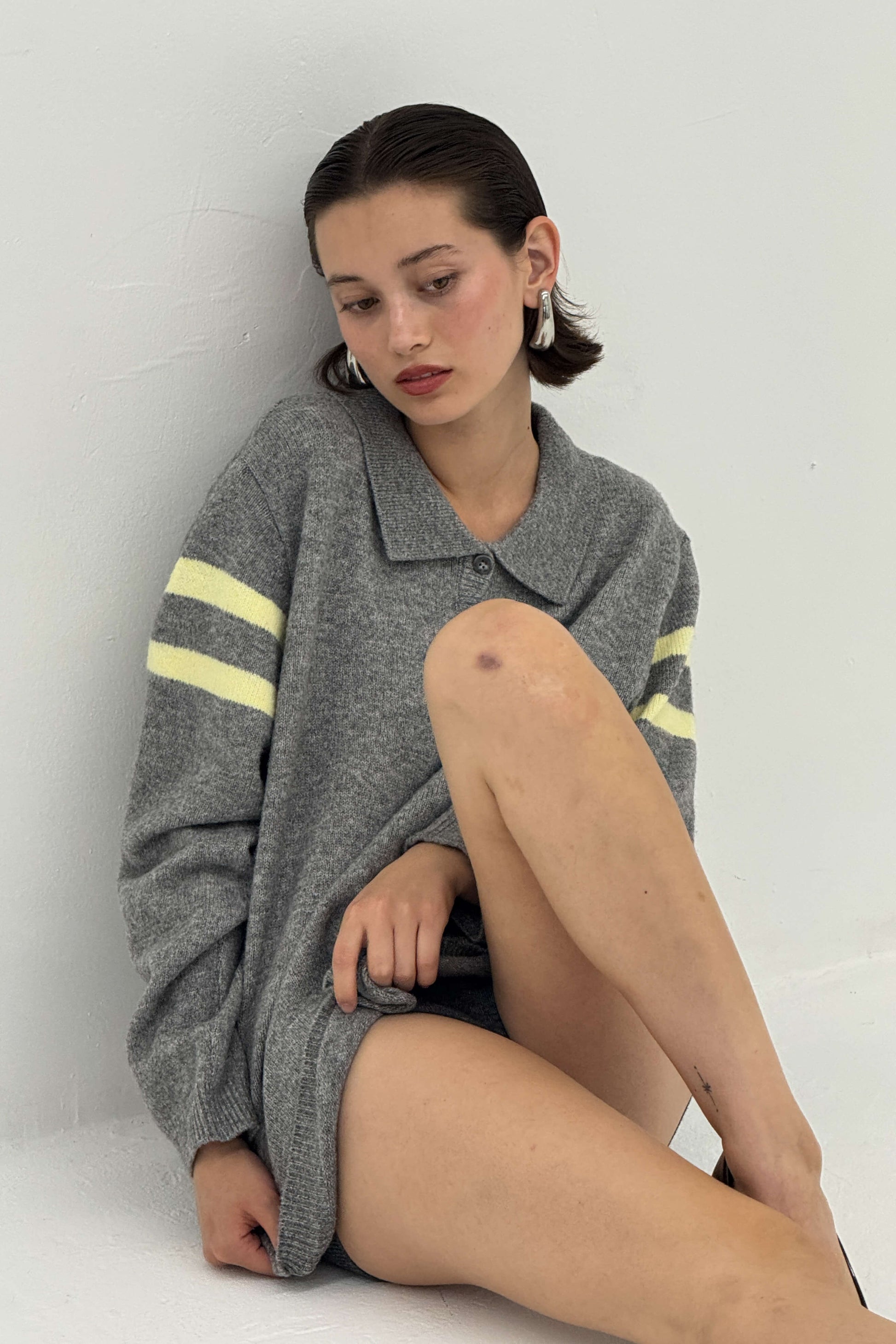 Woman wearing a gray sweater with yellow stripes sitting against a white wall.