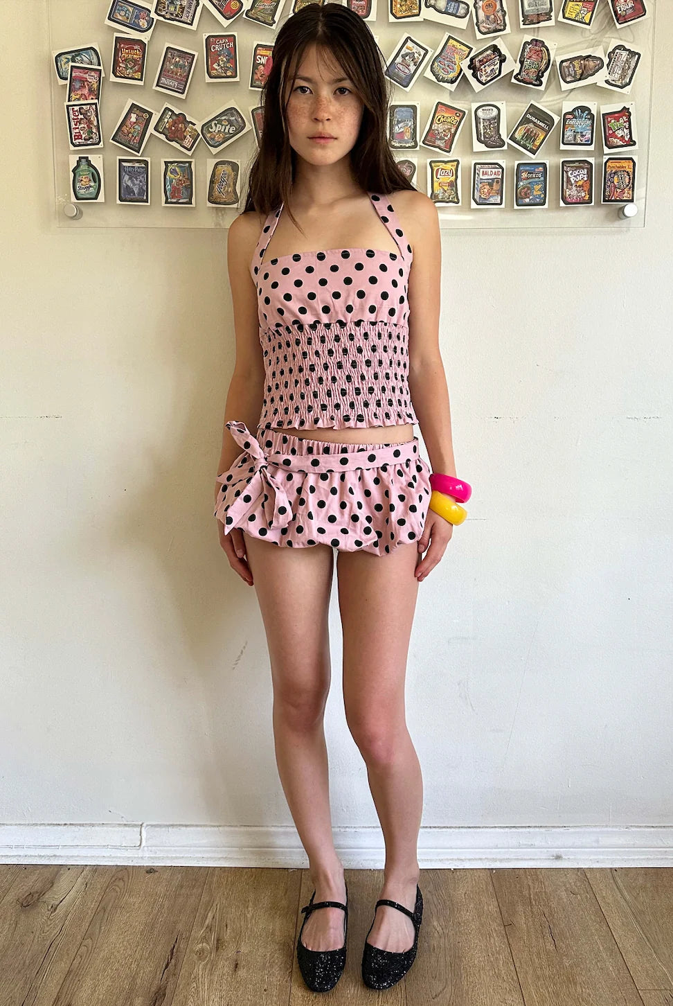 Woman wearing a pink polka dot dress with a colorful object in her hand, standing against a wall with decorative items.